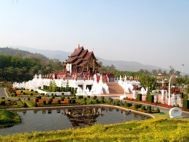 Le Pavillon Royal de style Lanna à Chiang Mai, en Thaïlande, entouré de jardins manucurés, avec un étang au premier plan et des montagnes en arrière-plan.