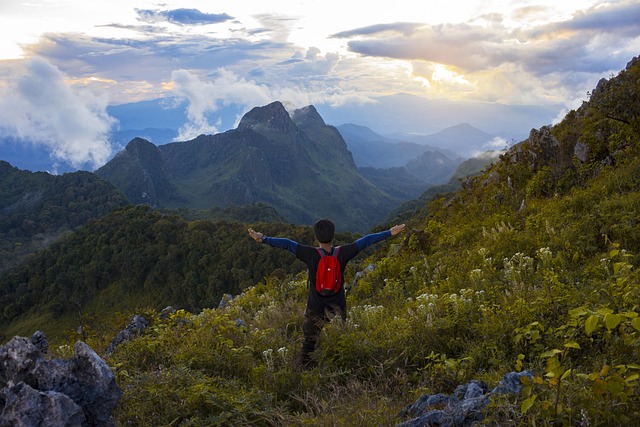 Un randonneur avec un sac à dos rouge, les bras ouverts, contemple une chaîne de montagnes verdoyantes en Thaïlande, illustrant la récompense de l'écotourisme.