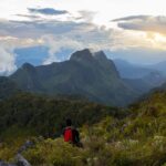 Randonneur avec un sac à dos rouge admirant la vue panoramique sur les sommets du Doi Chiang Dao au coucher du soleil.