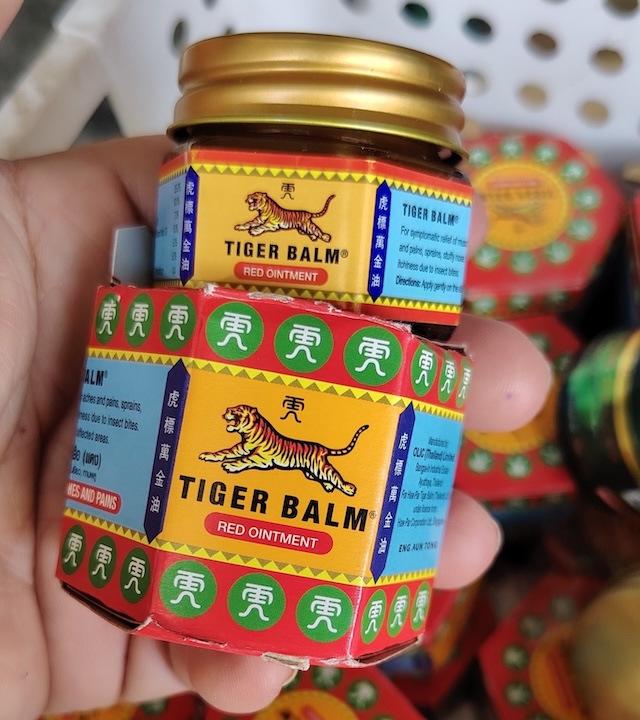 Gros plan sur un pot et une boîte de Baume du Tigre rouge (Red Ointment) tenus dans une main, un souvenir typique de Thaïlande.