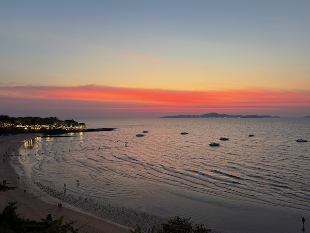 Vue de la plage de Pattaya en Thaïlande au crépuscule, avec un ciel aux couleurs chaudes, la mer et des bateaux au loin.