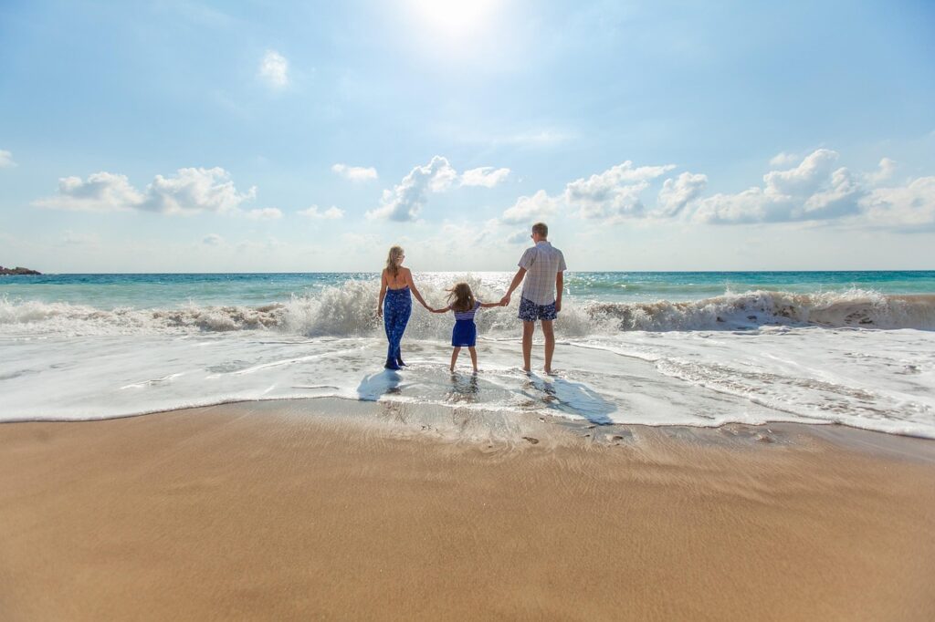 Famille composée de deux adultes et un enfant se tenant la main sur une plage en Thaïlande, face à la mer sous un ciel ensoleillé