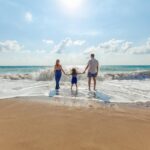 Famille composée de deux adultes et un enfant se tenant la main sur une plage en Thaïlande, face à la mer sous un ciel ensoleillé