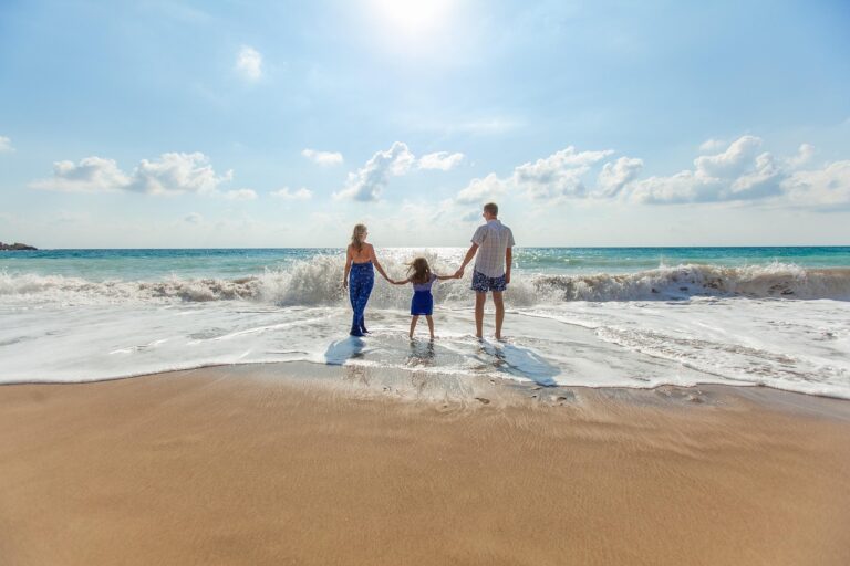 Famille composée de deux adultes et un enfant se tenant la main sur une plage en Thaïlande, face à la mer sous un ciel ensoleillé