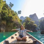 Voyageuse seule assise à l'avant d'un bateau en bois, les bras ouverts, souriant face au paysage du lac de Khao Sok en Thaïlande.