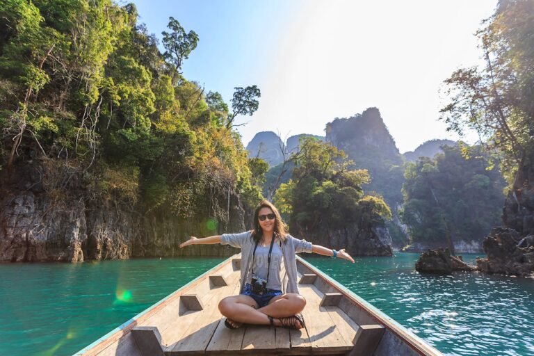 Voyageuse seule assise à l'avant d'un bateau en bois, les bras ouverts, souriant face au paysage du lac de Khao Sok en Thaïlande.
