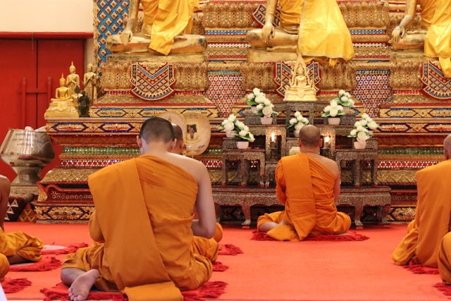 Vue de dos de moines bouddhistes en robe safran, assis en méditation sur un tapis rouge devant un autel doré dans un temple en Thaïlande.