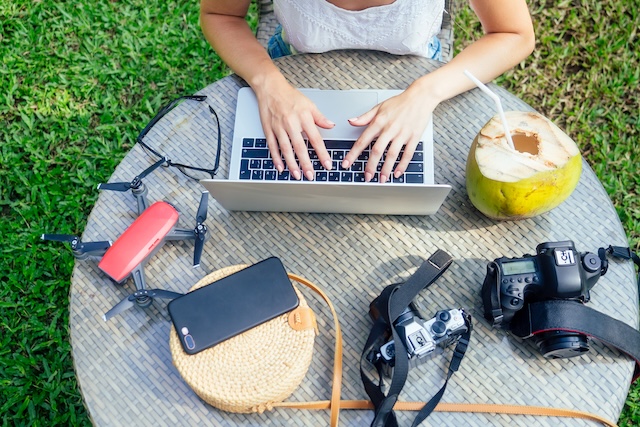 Une nomade digitale travaille sur son ordinateur en extérieur en Thaïlande, entourée de son matériel (drone, appareil photo) et d'une noix de coco, illustrant le style de vie des voyageurs avec un visa DTV.
