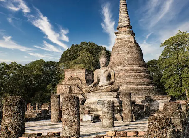Une statue de Bouddha en pierre méditant devant un grand stupa et les ruines du temple Wat Mahathat dans le parc historique de Sukhothai.