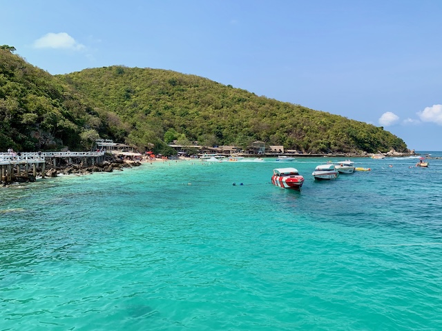 Vue de la plage de Tawaen sur l'île de Koh Larn, avec son eau turquoise, des bateaux rapides et une colline verdoyante sous un ciel bleu.