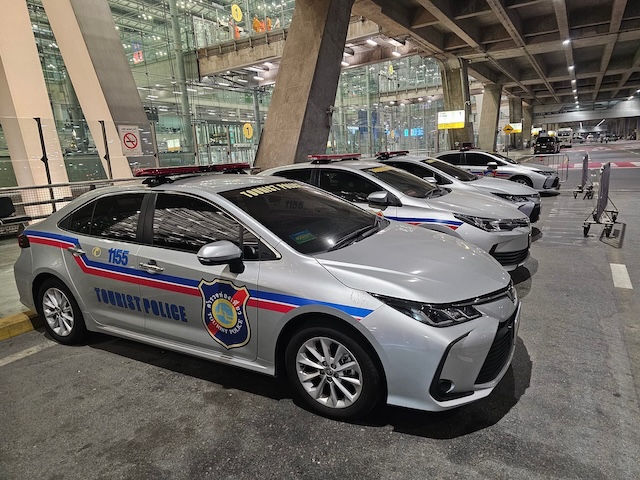 Plusieurs voitures de la Police Touristique de Thaïlande (Tourist Police) garées devant un aéroport, montrant le numéro d'urgence 1155.