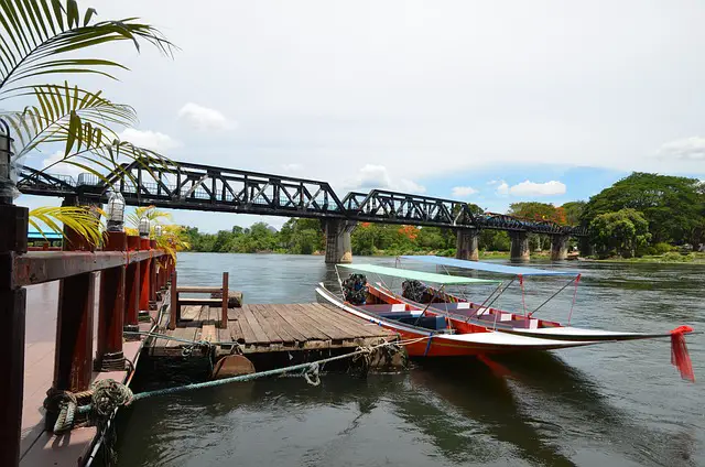 Vue du pont métallique de la rivière Kwaï à Kanchanaburi, avec des bateaux à longue queue amarrés à un ponton en bois au premier plan.