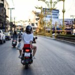 Femme vue de dos portant un casque en conduisant un scooter dans une rue en Thaïlande, illustrant la sécurité routière.