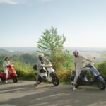 Trois voyageurs avec leurs casques, arrêtés sur leurs scooters sur une route de campagne en Thaïlande, admirant une vue sur la mer et la végétation.