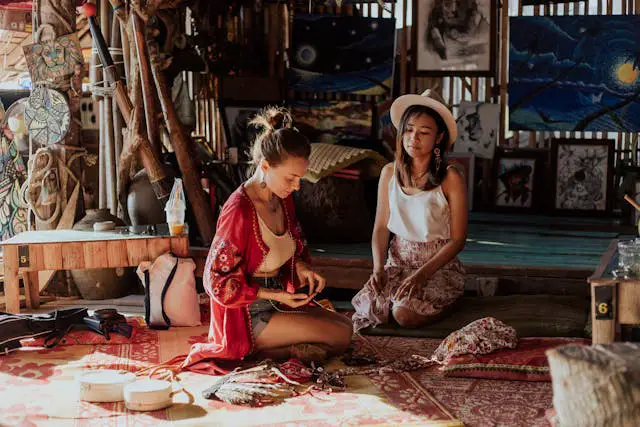 Deux jeunes femmes, une occidentale et une asiatique, assises sur des nattes dans un atelier d'artisanat en Thaïlande, examinant des bijoux locaux dans une ambiance calme et authentique.