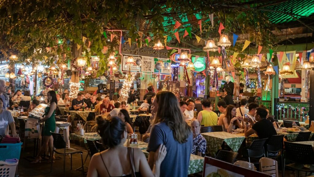 Touristes attablés la nuit dans un restaurant de rue animé à Khao San Road, Bangkok, avec des lumières et des guirlandes.
