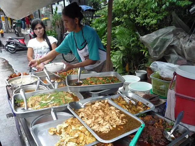 Vendeuse de street food thaïlandaise servant une cliente depuis son stand de rue rempli de plats locaux.