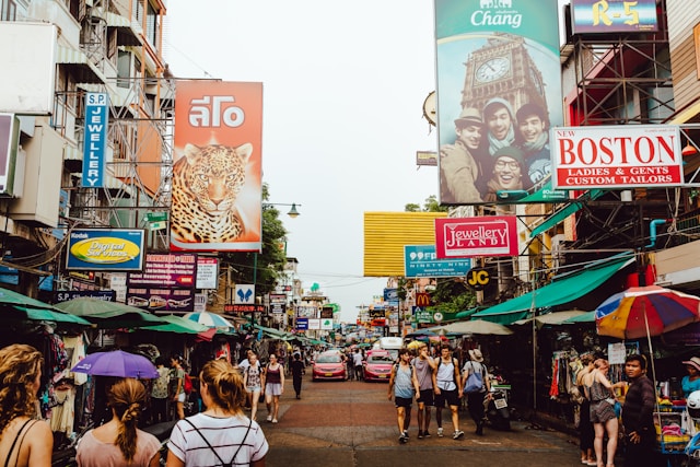 Vue de la rue Khao San Road en journée avec ses nombreux panneaux publicitaires, ses étals de marché et les touristes qui se promènent.