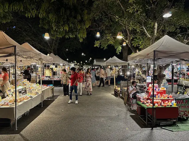 Allée d'un marché de nuit animé en Thaïlande, avec des stands illuminés sous des arbres et des visiteurs qui se promènent.