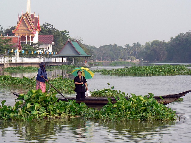 Deux femmes sur une barque en bois traditionnelle naviguant sur un fleuve en Thaïlande, avec un temple thaïlandais en arrière-plan.