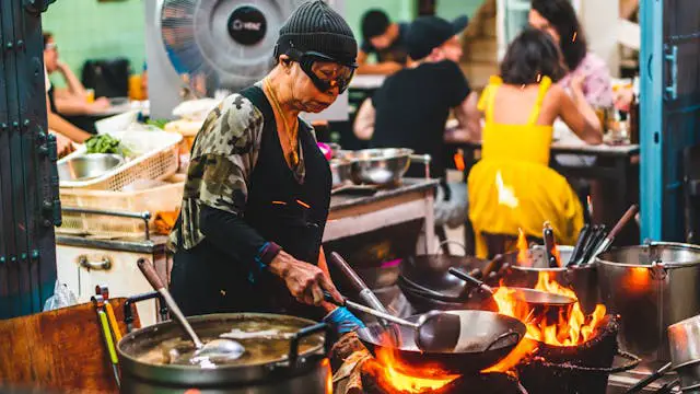 a célèbre cheffe de street food thaïlandaise Jay Fai cuisinant au wok sur un feu vif, illustrant la sécurité et l'hygiène de la cuisine de rue en Thaïlande.