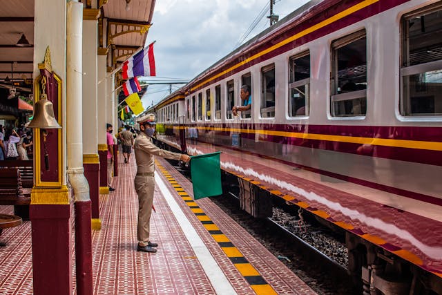 Chef de gare en uniforme agitant un drapeau vert devant un train thaïlandais à quai, signalant le départ.