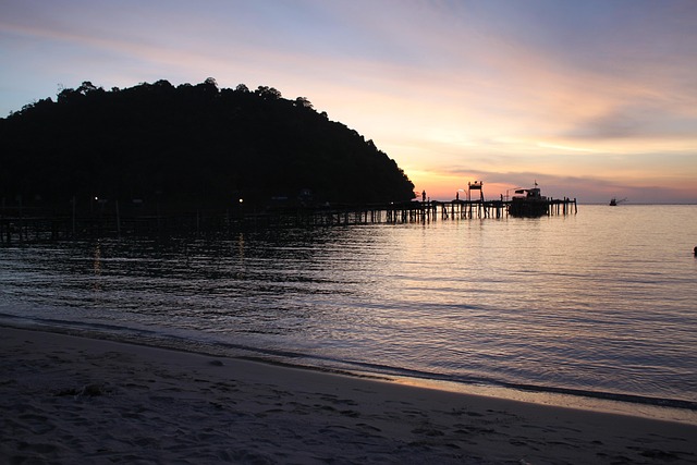 Coucher de soleil paisible sur une plage de Koh Kood avec un long ponton en bois s'avançant dans la mer et des bateaux de pêche au loin.