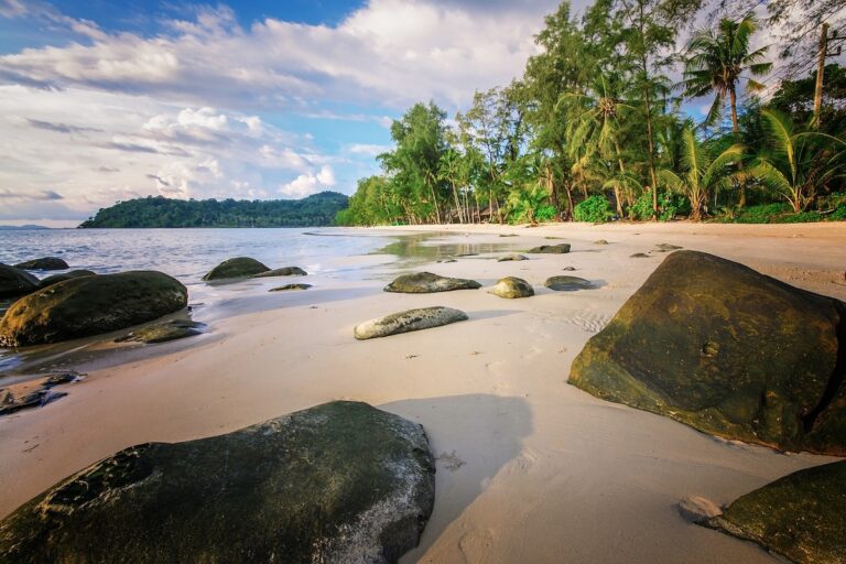 Paysage paradisiaque d'une plage sauvage à Koh Kood en Thaïlande : sable fin, gros rochers au premier plan et cocotiers bordant une mer calme au coucher du soleil.