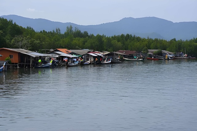 Maisons sur pilotis et bateaux de pêche alignés le long d’un village de pêcheurs près de Khanom, devant une mangrove et des collines