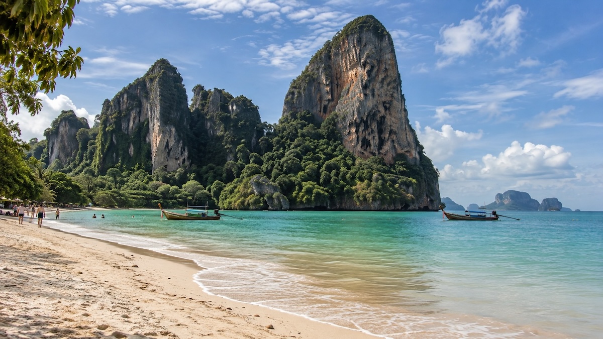 Plage de sable bordée de falaises calcaires à Krabi, avec des bateaux traditionnels thaïlandais sur l'eau turquoise.